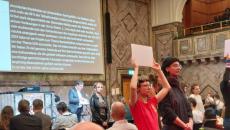 Scène de la manifestation à Zurich. On y voit de dos les auditrices et auditeurs, avec à l’arrière-plan, un conférencier devant un visuel projeté à l’écran et au premier plan, deux étudiantes qui tournent le dos à l’orateur et arborent une pancarte avec un slogan.