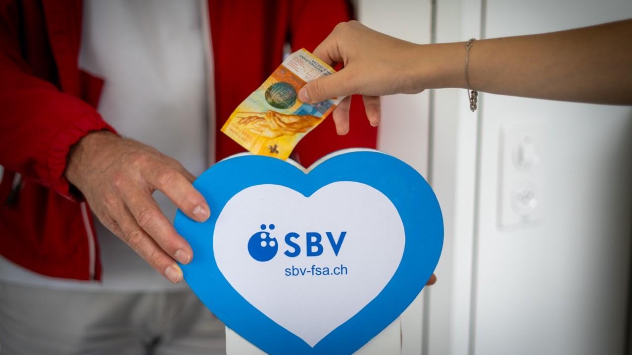 Symbolbild in Nahaufnhame: Ein Mann hält an der Türe eine Kasse mit dem Aufdruck "SBV" in der Hand. Eine andere Person wirft einen 10-Franken-Schein hinein.