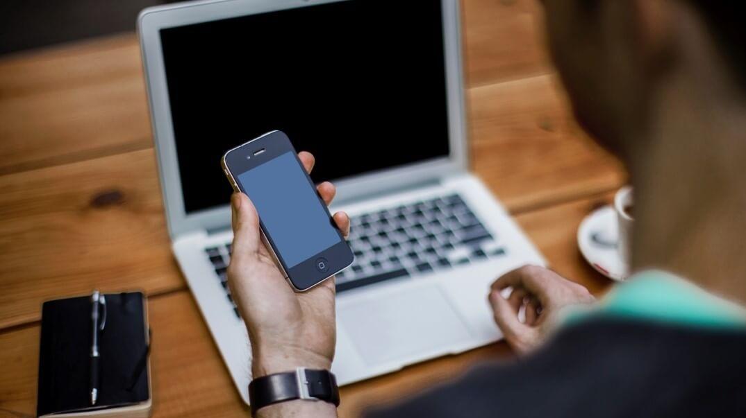 Symbolbild: Ein Mann sitzt vor einem Laptop und schaut dabei auf sein Smartphone, dass er in der Hand hält. Beide Displays sind schwarz.
