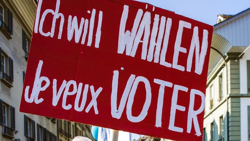 Gros plan sur une pancarte de manifestation lors d'une précédente manifestation. Sur fond rouge, on peut lire en blanc : "Ich will WÄHLEN - Je veux VOTER".