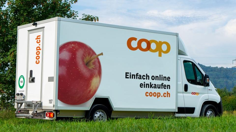 Ein weisser Coop-Lieferwagen fährt durch die Landschaft. Auf dem Lieferwagen steht: "Coop; Einfach online einkaufen; coop.ch". Links daneben ist ein grosser roter Apfel aufgedruckt.