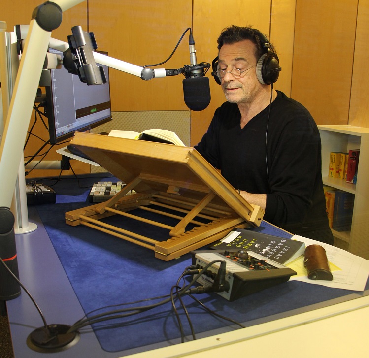 Das Bild zeigt einen Mann mit Brille und aufgesetzten Kopfhörern in einem schalldichten Raum. Er sitzt vor einem Lesepult mit aufgeschlagenem Buch und spricht in ein grosses Mikrofon. 