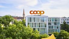 Photo du siège social de Coop à Bâle. Sur le toit, en grande lettre, l’inscription «Coop». A l’arrière plan, à gauche, on voit une église et au premier plan, des arbres en feuilles. 