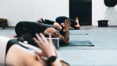 Quatre personnes côte à côte dans une salle, agenouillées sur le sol. Elles font toutes le même exercice de yoga, la tête baissée contre le sol et les bras avec les mains croisées pliés vers le haut. 