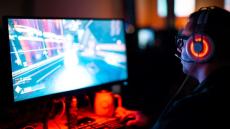 Unscharf zu erkennender PC-Bildschirm, auf dem ein Game läuft. Davor sitzt ein Mann mit leuchtendem Headset im Halbdunkel. 