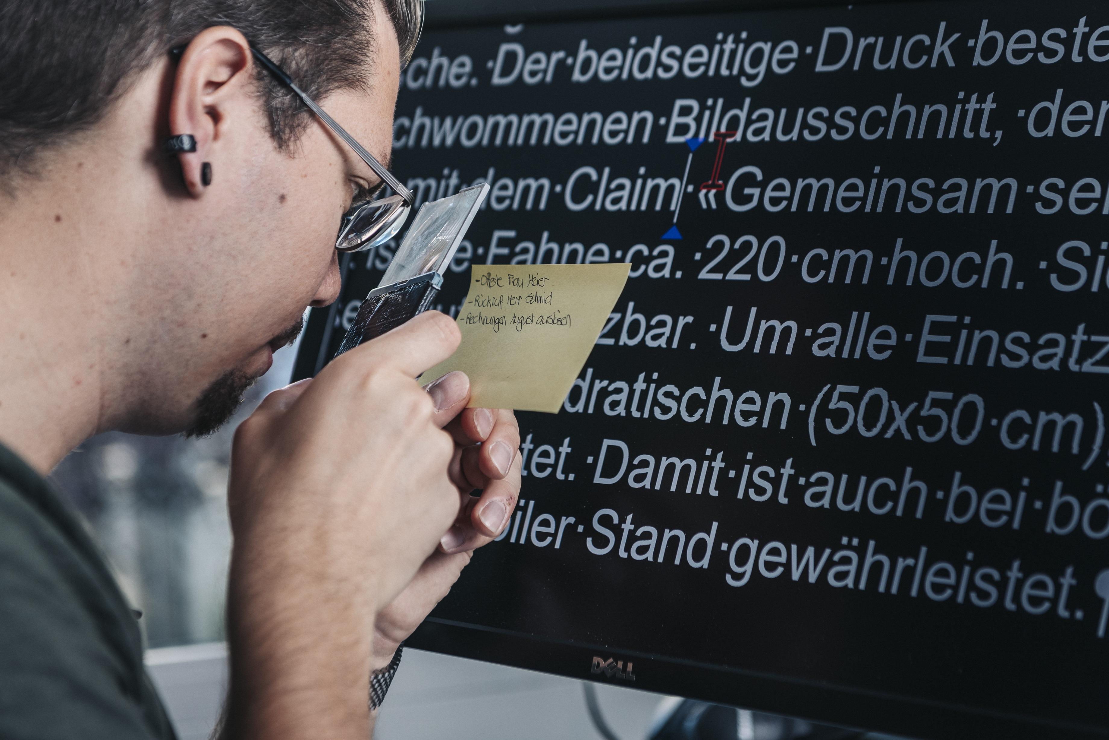 sehbehinderter Mann vor PC Bildschirm mit Vergrösserungsoftware und Handlupe für kleine Notizen