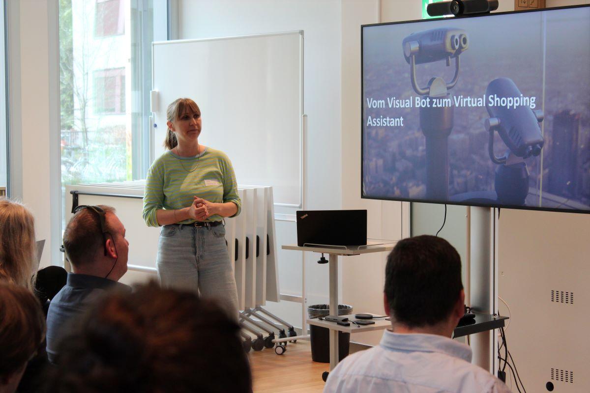 Une femme présente le projet informatique au public, dont quatre personnes sont visibles. L'écran affiche "Du Visual Bot à l'assistant virtuel d'achat".