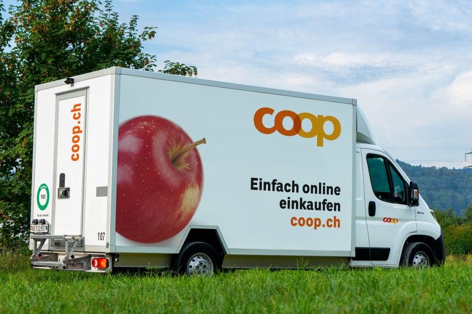Ein weisser Coop-Lieferwagen fährt durch die Landschaft. Auf dem Lieferwagen steht: "Coop; Einfach online einkaufen; coop.ch". Links daneben ist ein grosser roter Apfel aufgedruckt.