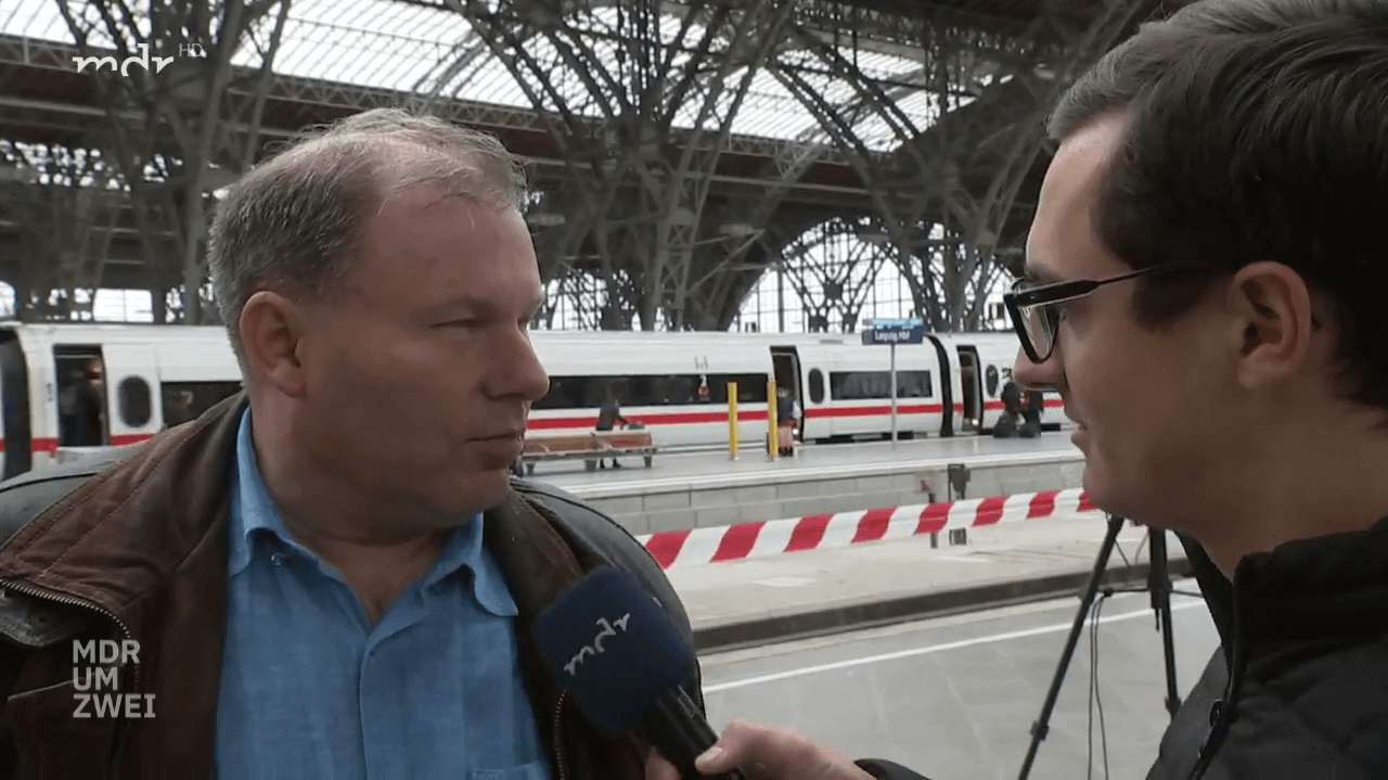 Ausschnitt aus einem Fernsehinterview: Ein Reporter interviewt einen SBV-Mitarbeiter an den Gleisen eines grossen Bahnhofshalle. Im Hintergrund steht ein deutscher ICE-Zug.